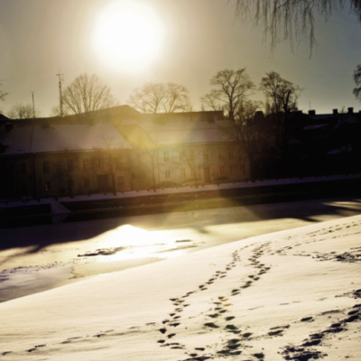 Spår i snön fram ett fruset vatten och ett äldre hus. Solen går ner ovanför huset.