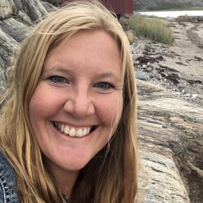 Den leende forskaren Sara Blanck i närbild med klippor och vatten i bakgrunden.