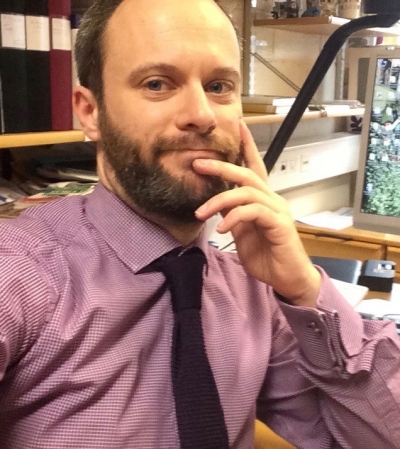 Selfie på Kent Fredholm med skägg, rödrutig skjorta och svart slips sittande på sitt kontor.