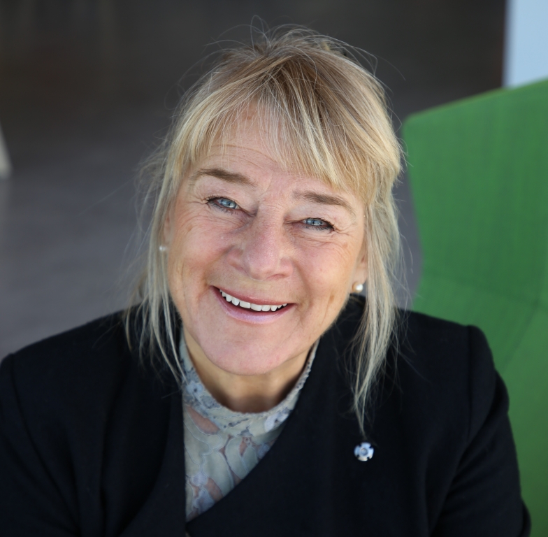 Blond medelålders kvinna i svart kavaj med pärlörhängen som ler mot kameran. Ansiktet är i närbild och bakgrunden är suddig i grönt och grått.