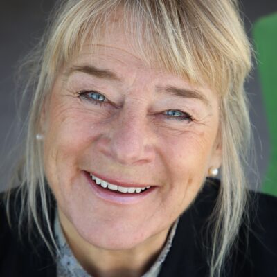Blond medelålders kvinna i svart kavaj med pärlörhängen som ler mot kameran. Ansiktet är i närbild och bakgrunden är suddig i grönt och grått.